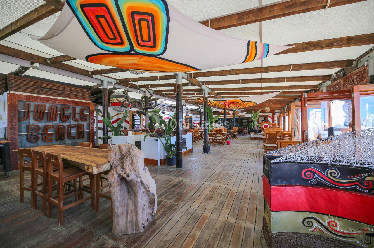 Terrasse en bois d'un restaurant décoré de toiles colorées et de mobilier en bois, avec des plantes en arrière-plan.