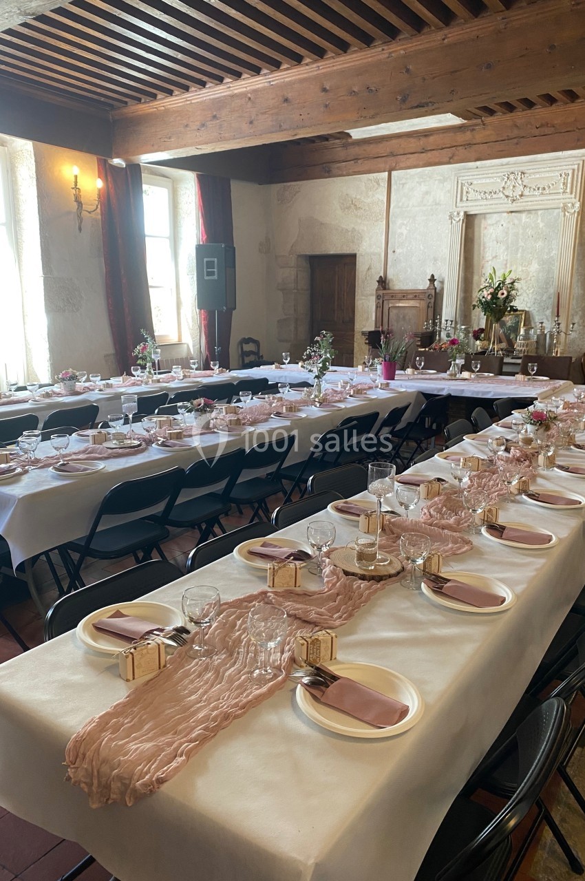 Salle de réception avec de longues tables dressées, nappes blanches, chemins de table roses et décoration florale.