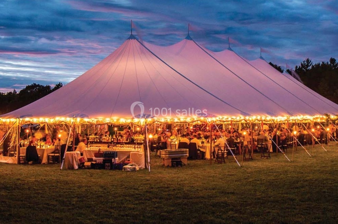 Grande tente éclairée par des guirlandes lumineuses, abritant un événement en soirée sur une pelouse.