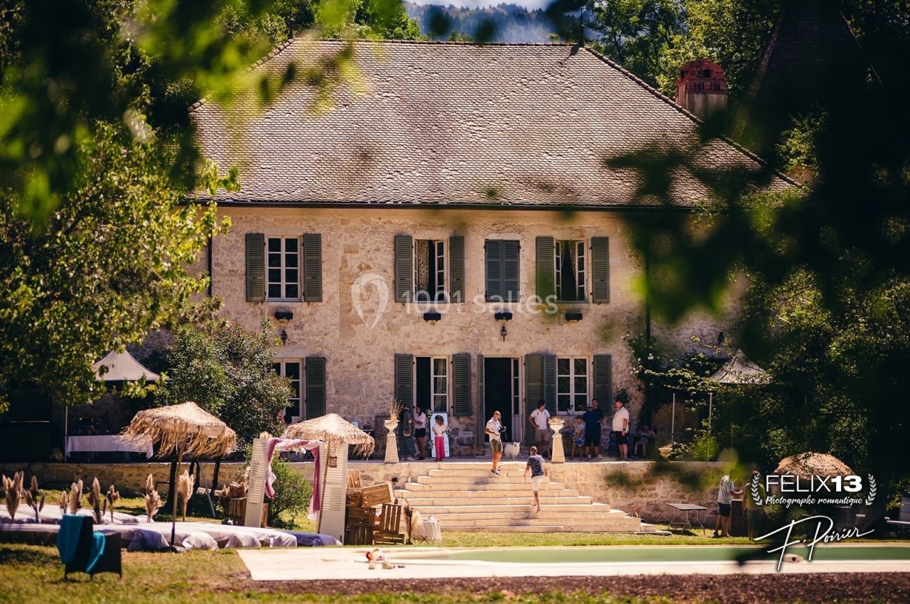 Grande maison en pierre avec volets verts, entourée de verdure, des personnes rassemblées devant l'entrée.