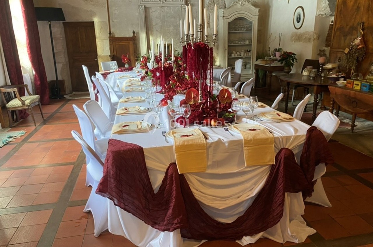 Table de banquet élégamment dressée avec nappes blanches, décorations rouges et chandeliers dans une salle rustique.