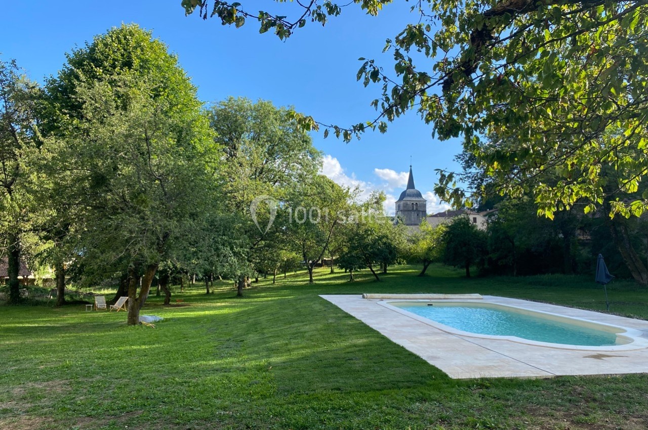 Jardin verdoyant avec piscine rectangulaire, arbres ombragés et clocher d'église visible en arrière-plan.
