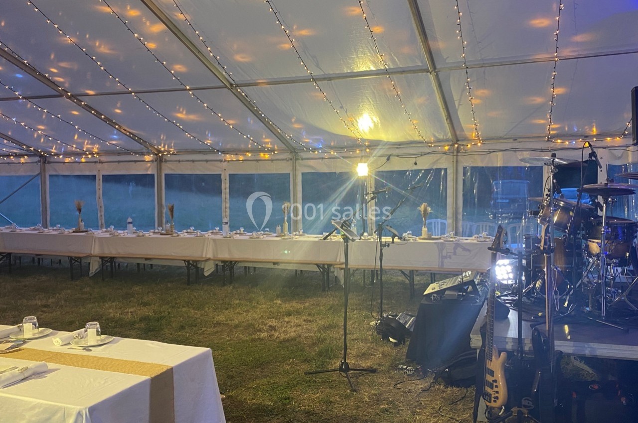 Intérieur d'une tente éclairée avec des guirlandes lumineuses, tables dressées et scène équipée d'instruments de musique.