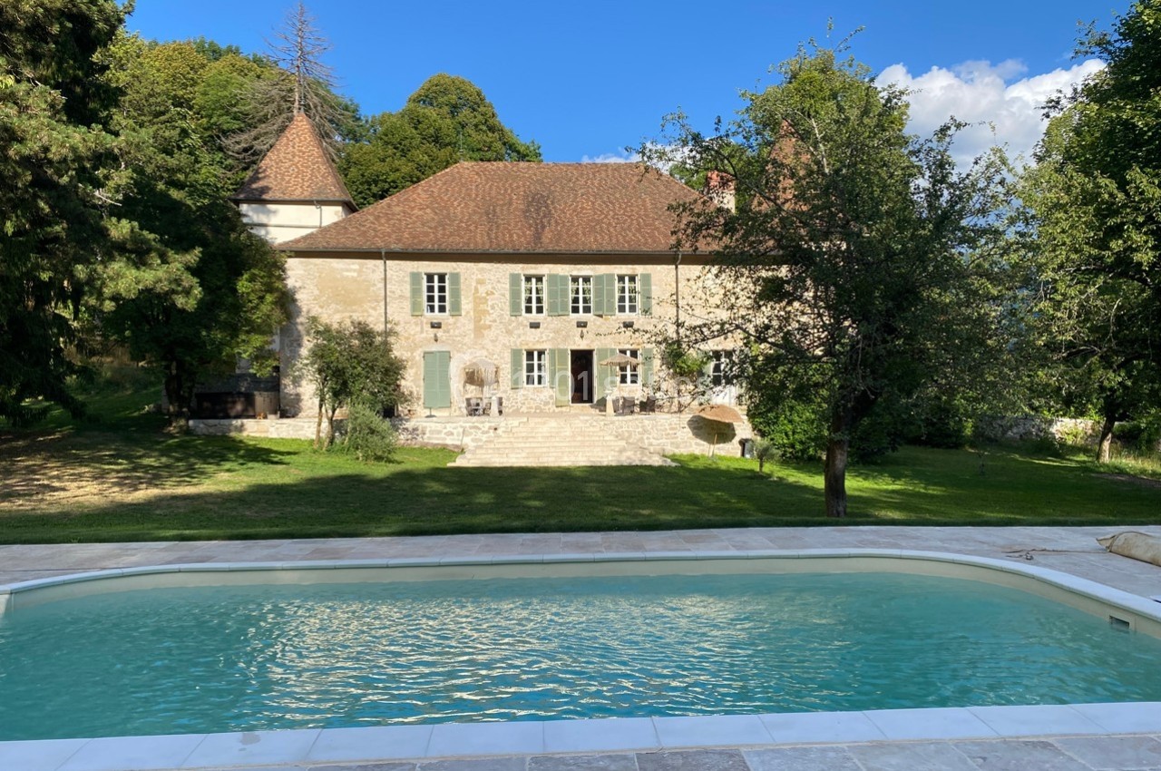 Façade d'une maison en pierre avec toit en tuiles, entourée d'arbres, vue depuis une piscine.