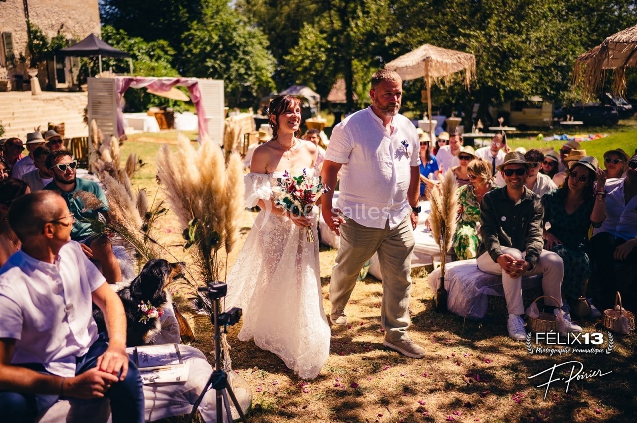 Une mariée en robe blanche marche dans une allée extérieure, accompagnée d'un homme, entourée d'invités assis.
