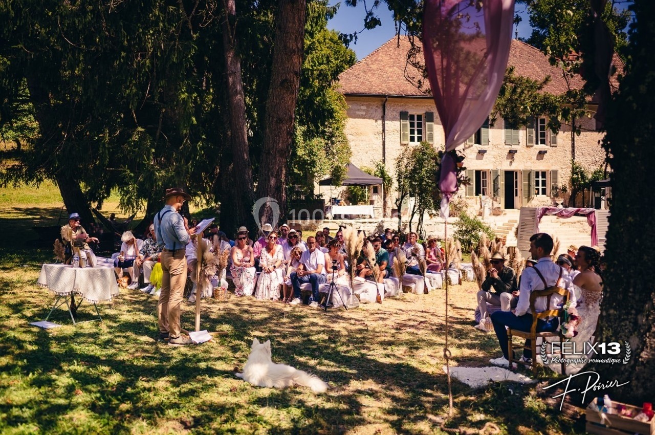 Cérémonie en plein air avec des invités assis sous des arbres, devant une maison en pierre.