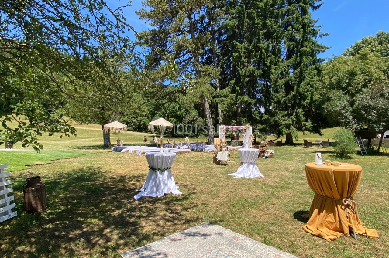 Espace extérieur aménagé pour un événement avec tables hautes, chaises, tapis et décorations sous des arbres.