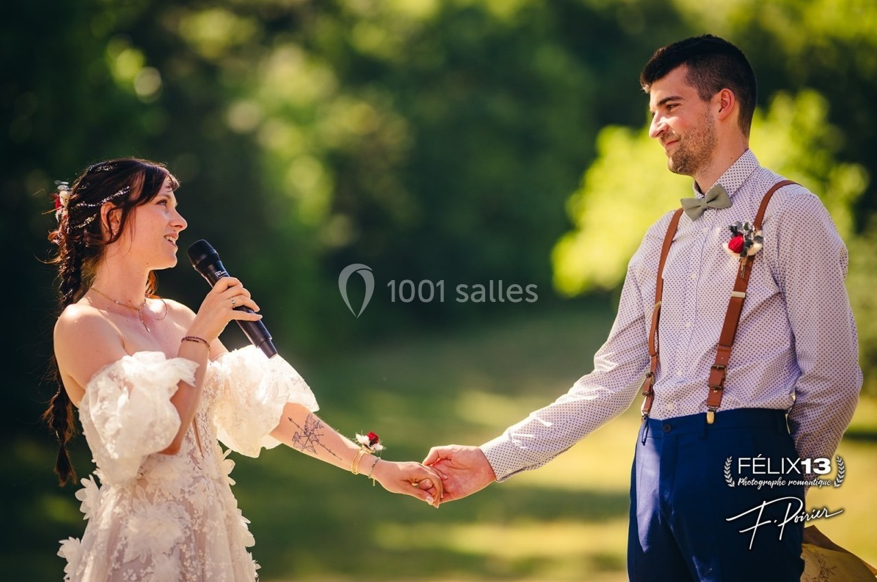 Une mariée tenant un micro s'adresse à son époux, main dans la main, dans un cadre extérieur verdoyant.