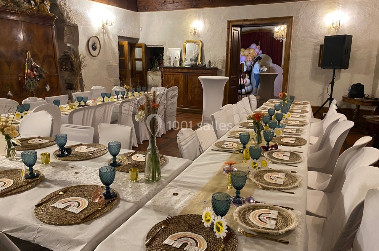 Salle décorée pour un événement avec longues tables dressées, nappes blanches, assiettes en osier et verres colorés.