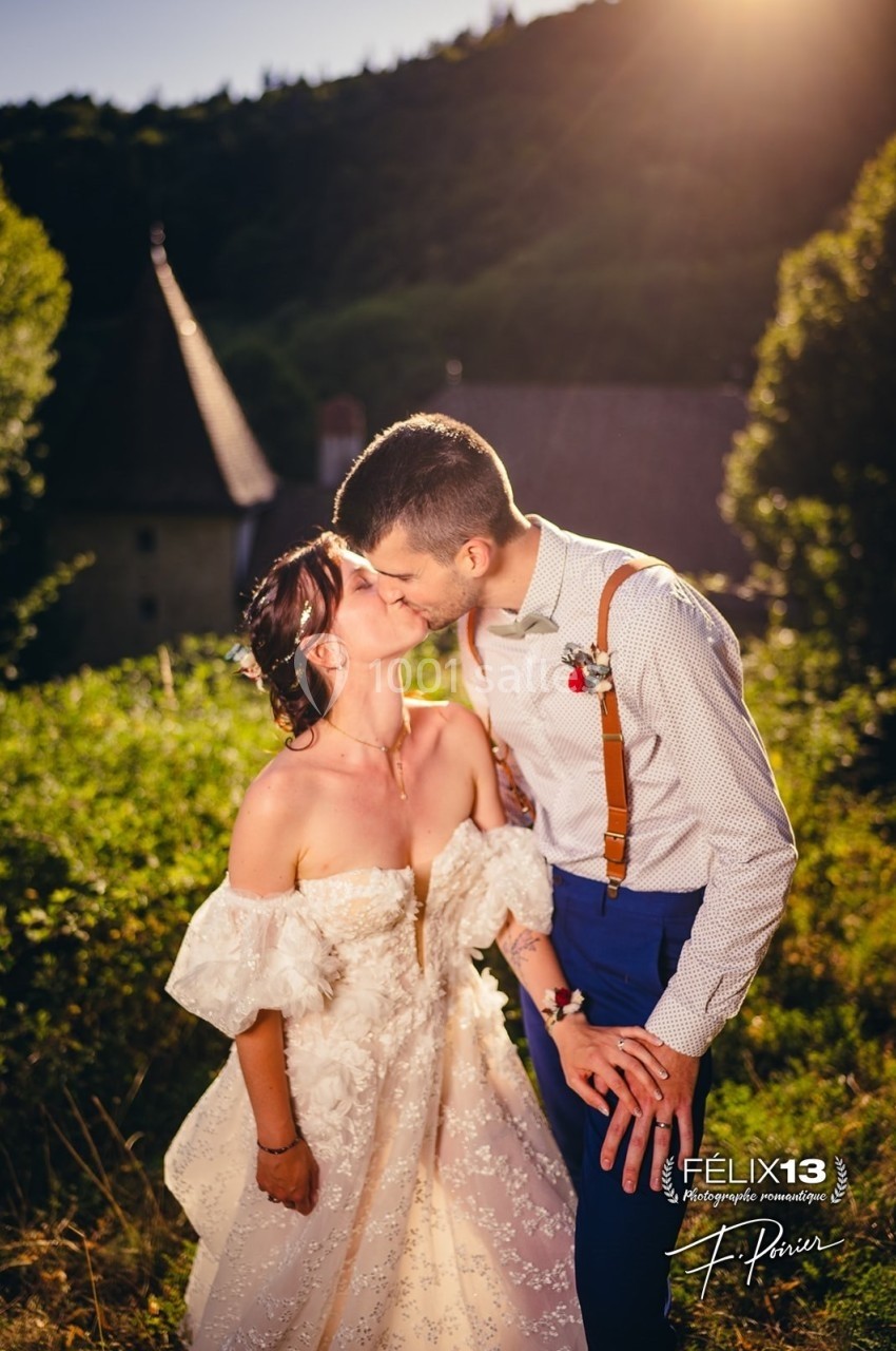 Un couple en tenue de mariage s'embrasse dans un cadre naturel ensoleillé avec des bâtiments en arrière-plan.