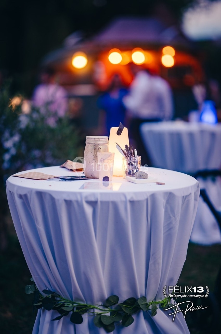Table haute décorée avec une bougie, un pot en verre et des couverts, dans un espace extérieur éclairé en soirée.