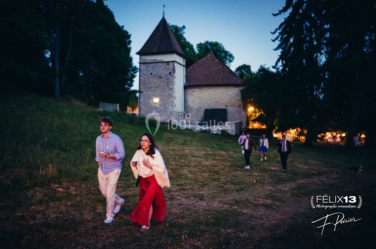 Un couple marche sur une pelouse au crépuscule devant un bâtiment en pierre éclairé, suivi par d'autres personnes.