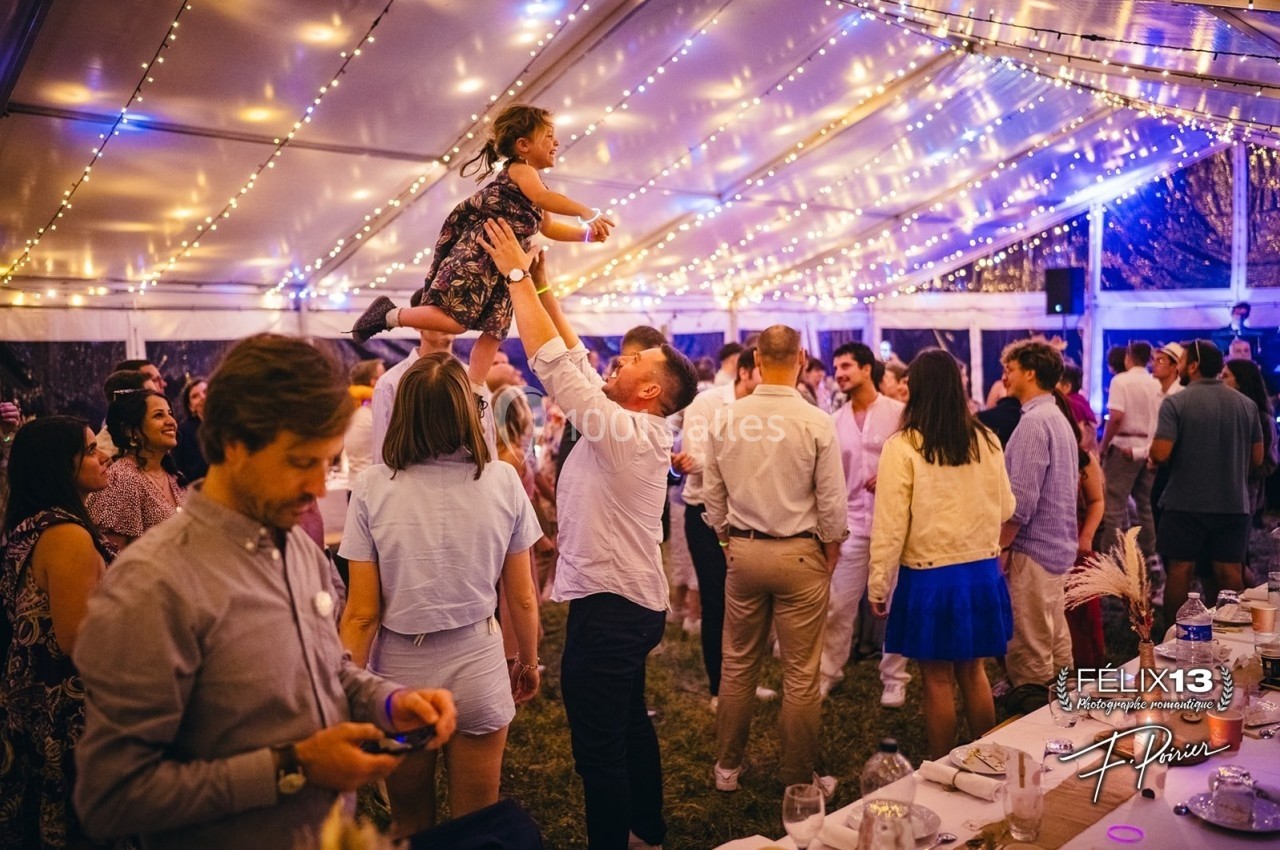 Un homme soulève une enfant dans ses bras au milieu d'une fête sous une tente illuminée de guirlandes.