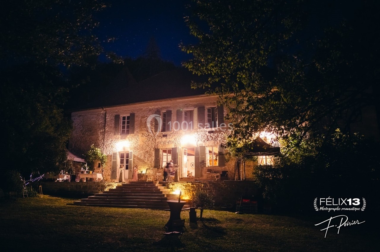 Façade d'une maison en pierre éclairée la nuit, entourée d'arbres et d'un jardin avec des lumières tamisées.