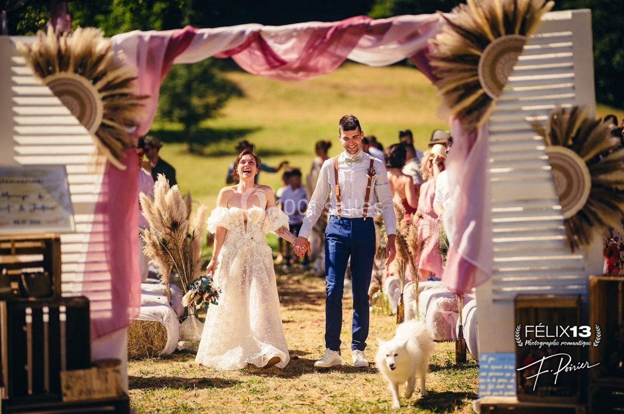 Un couple souriant marche main dans la main lors d'une cérémonie de mariage en plein air, entouré d'invités.