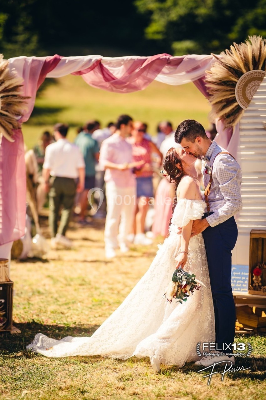 Un couple en tenue de mariage s'embrasse sous une arche décorée, avec des invités flous en arrière-plan.