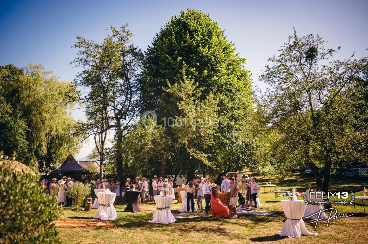 Groupe de personnes rassemblées dans un jardin lors d'un événement en plein air, entourées de tables hautes et d'arbres.