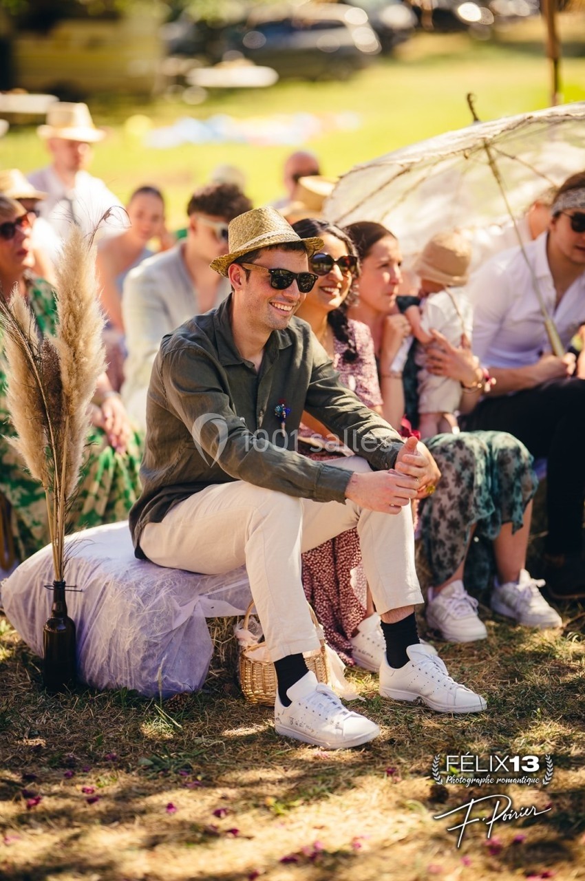 Un homme souriant, assis en extérieur lors d'un événement, entouré de personnes sous un soleil éclatant.