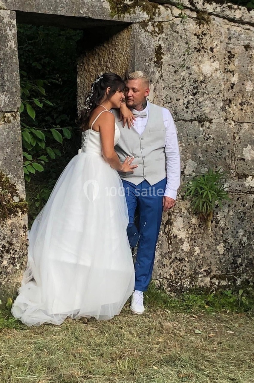 Un couple en tenue de mariage pose devant un mur en pierre dans un cadre naturel verdoyant.