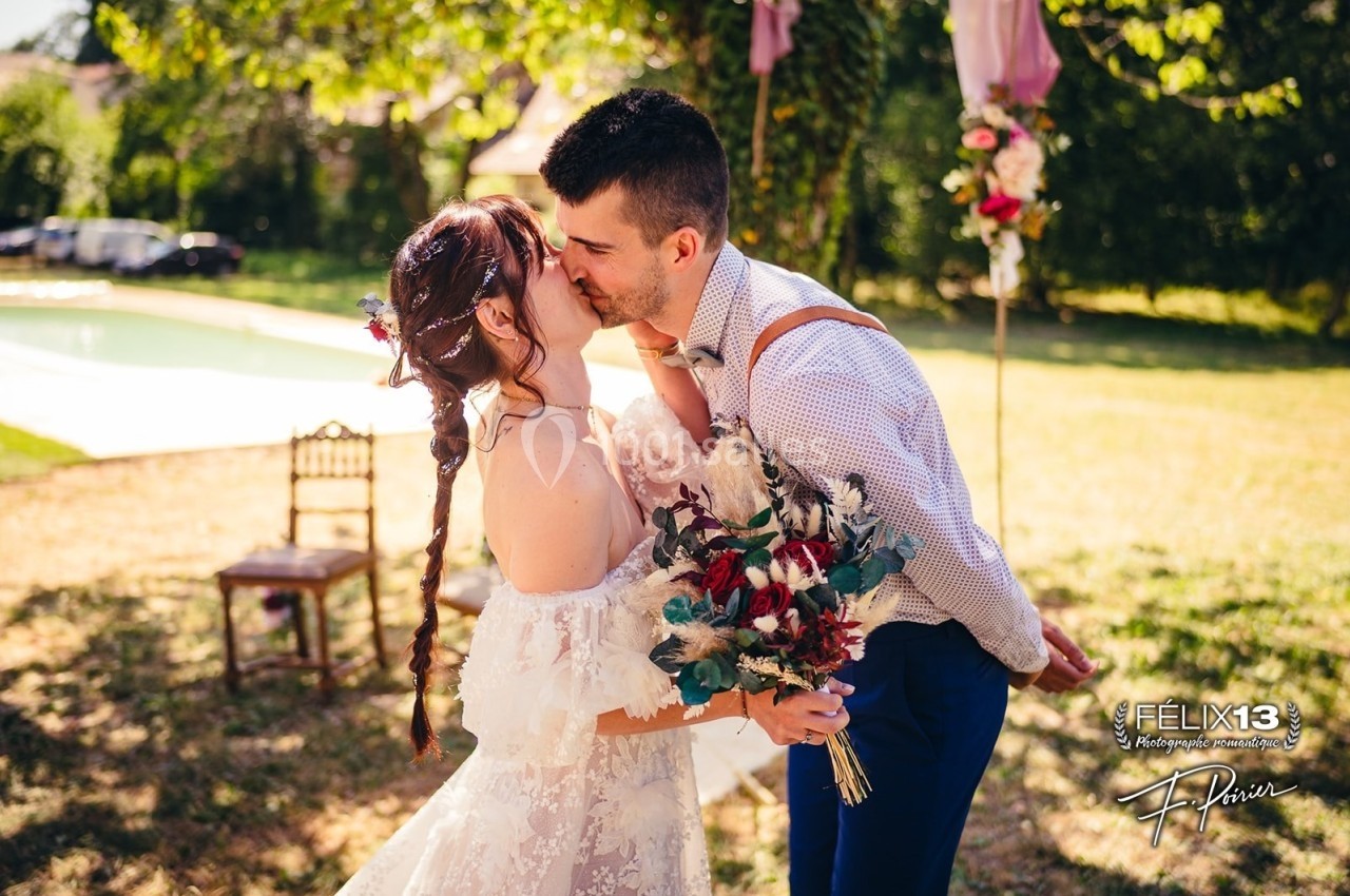 Un couple s'embrasse lors d'un mariage en extérieur, près d'une piscine, avec un bouquet de fleurs à la main.