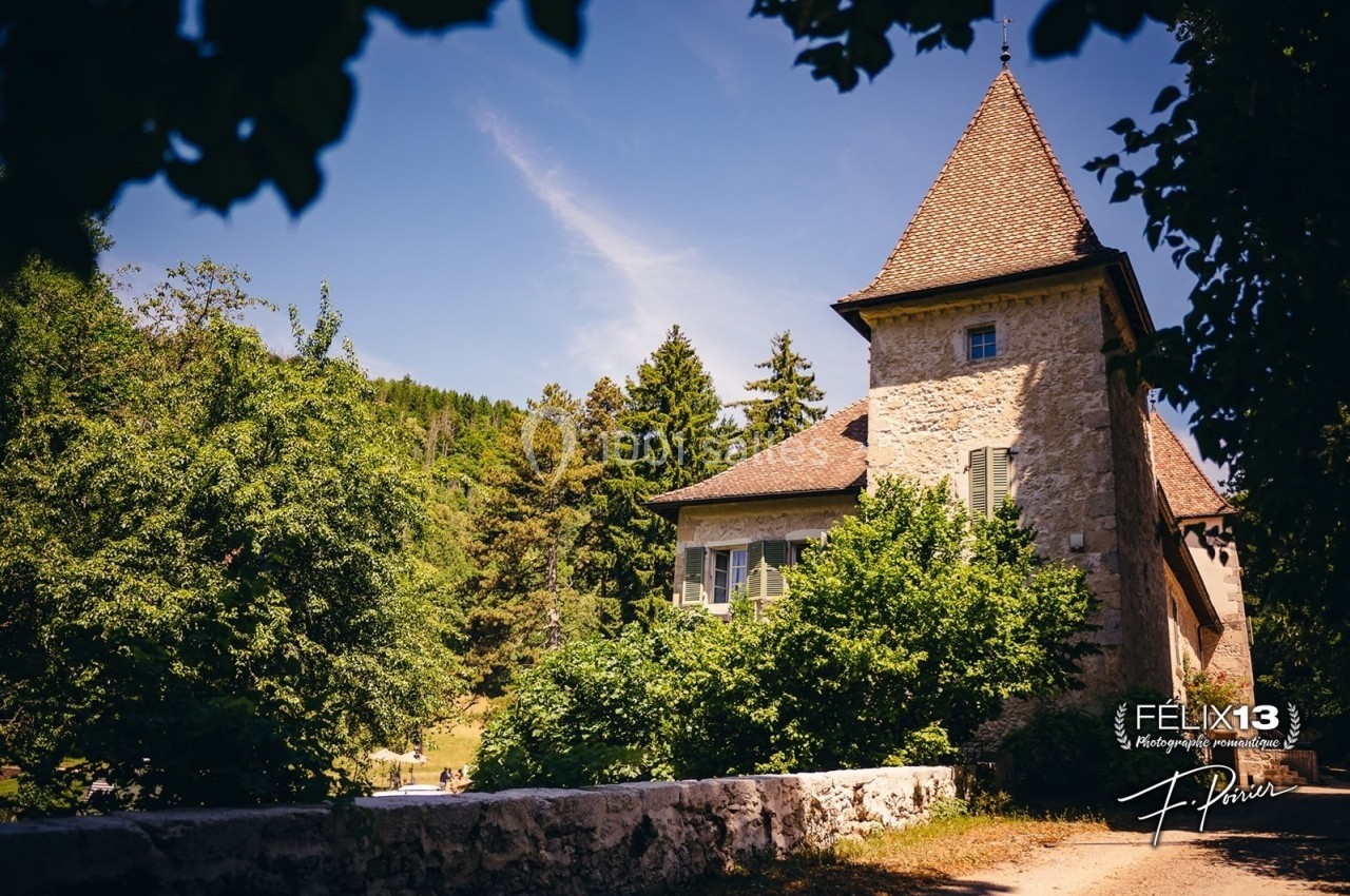 Maison en pierre avec toit en tuiles, entourée de verdure et d'arbres, sous un ciel bleu ensoleillé.
