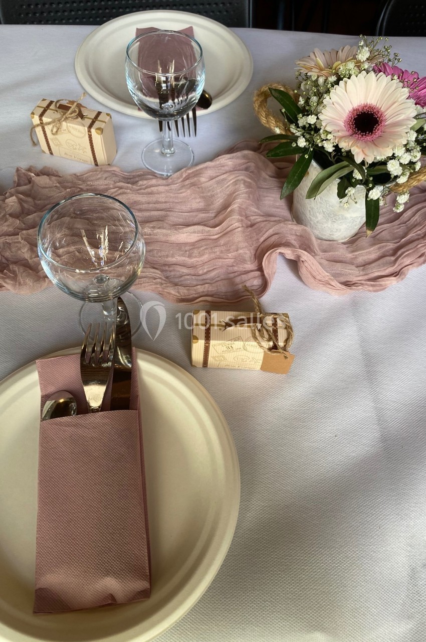 Table dressée avec vaisselle blanche, serviettes roses, petits cadeaux, chemin de table rose et bouquet de fleurs.