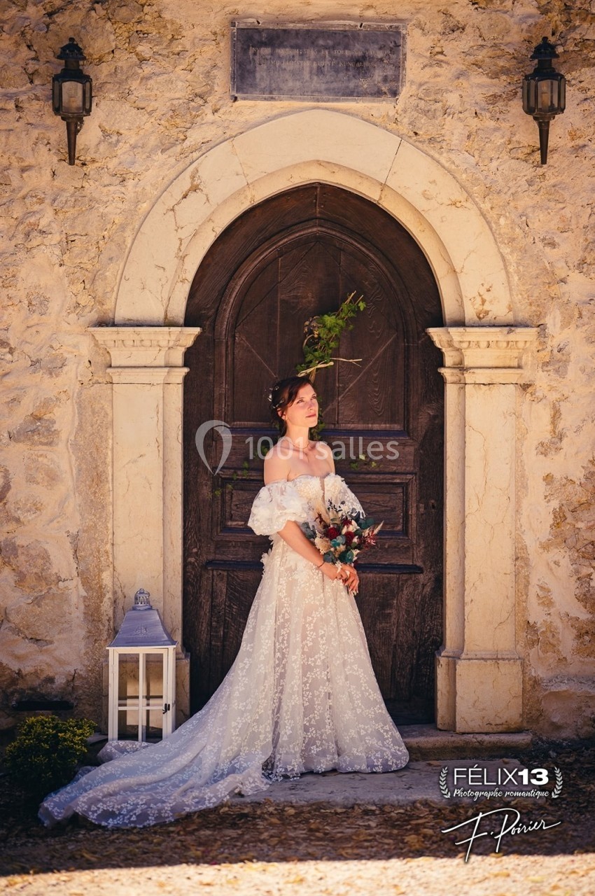 Femme en robe de mariée blanche tenant un bouquet, debout devant une porte en bois encadrée de pierre.