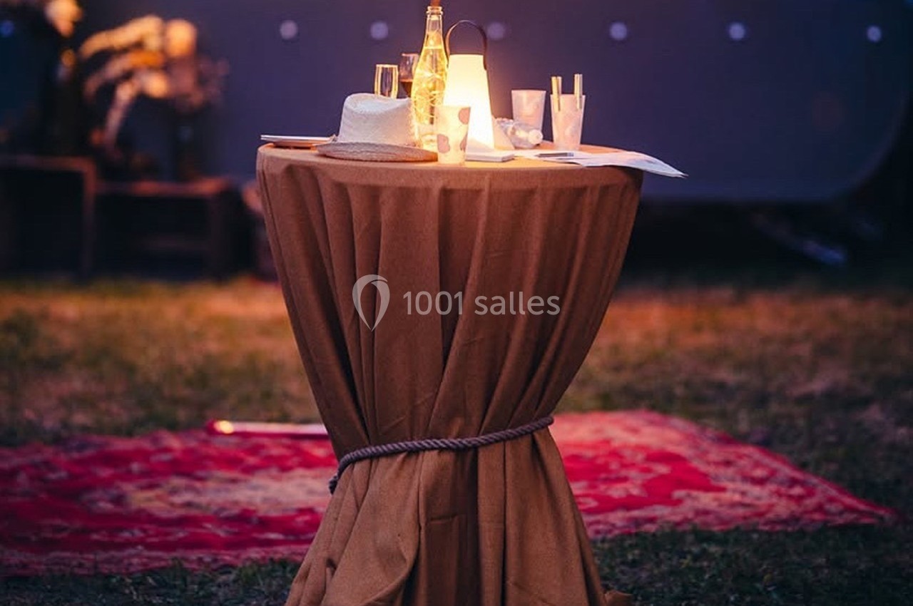 Table ronde couverte d'une nappe, décorée avec une lampe, un chapeau, des verres et une bouteille, en extérieur le soir.