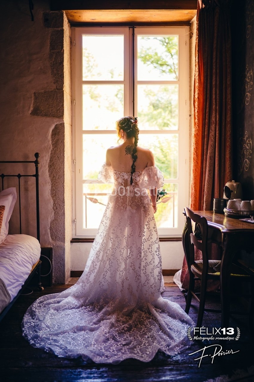 Une femme en robe de mariée en dentelle regarde par une fenêtre lumineuse dans une chambre au style rustique.