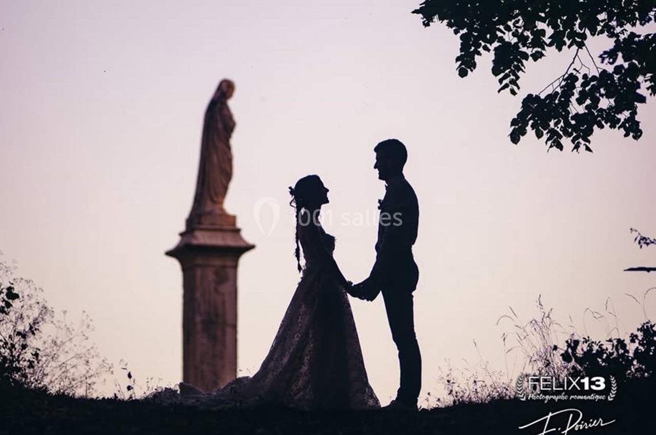 Un couple en tenue de mariage se tient la main devant une statue, au coucher du soleil.