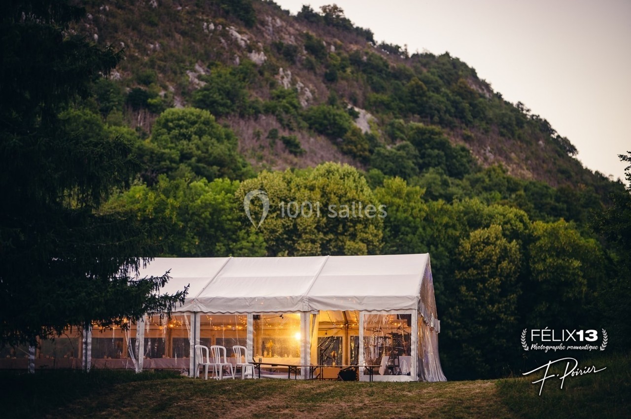 Grande tente blanche éclairée de l'intérieur, installée sur une pelouse au pied d'une colline boisée.