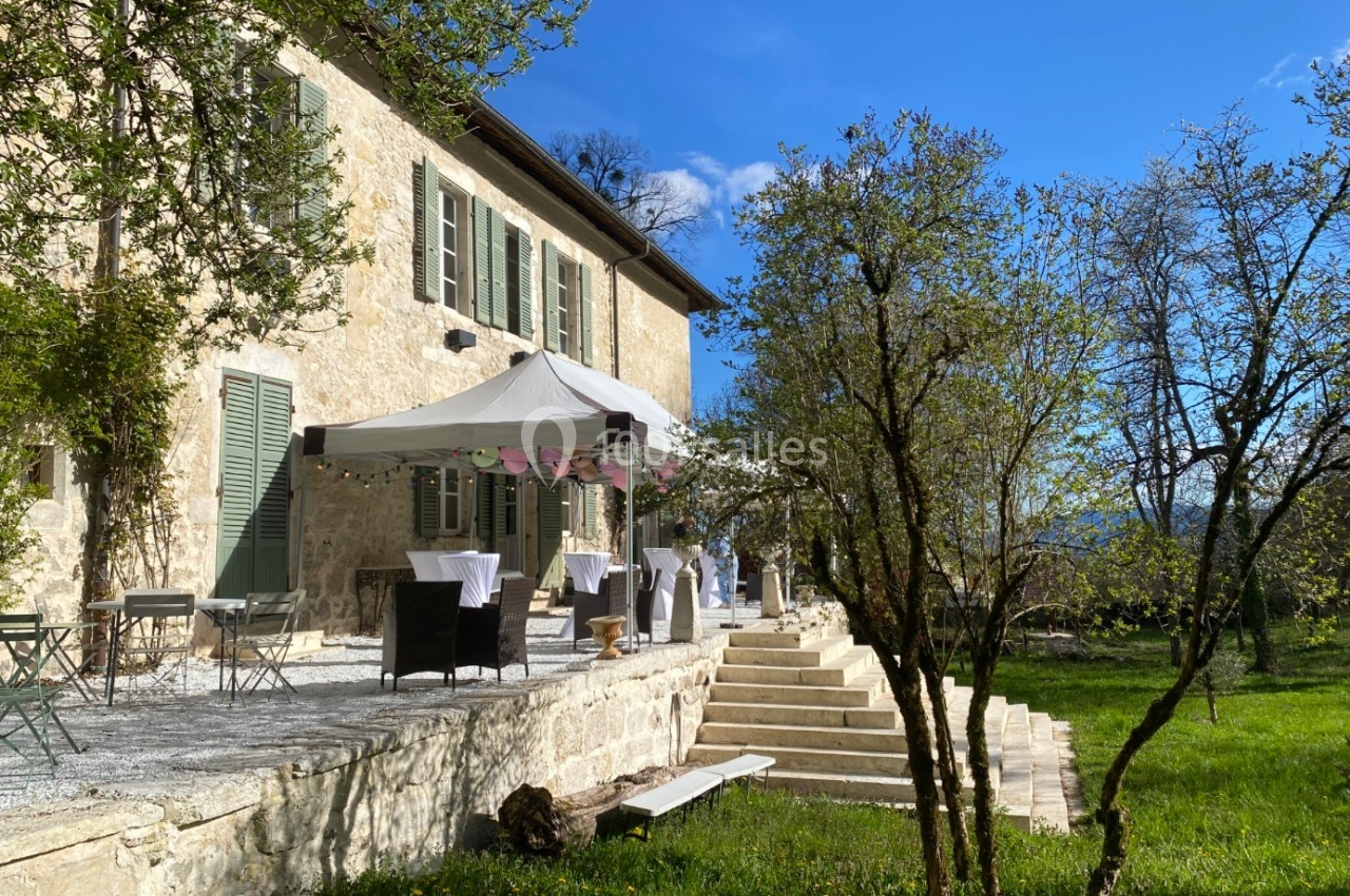 Terrasse d'une maison en pierre avec des tables dressées sous un barnum, entourée d'arbres et d'un jardin verdoyant.