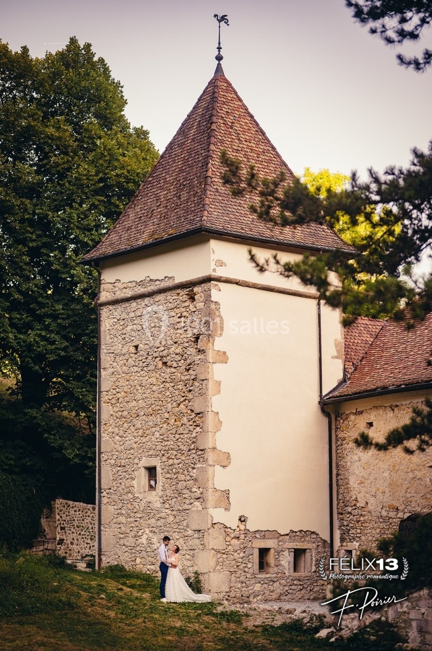 Tour en pierre ancienne avec un toit en tuiles, entourée de verdure, un couple en tenue de mariage au pied.