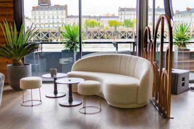 Terrasse avec tables et chaises en bord de rivière, entourée de plantes vertes et vue sur des bâtiments et un pont.