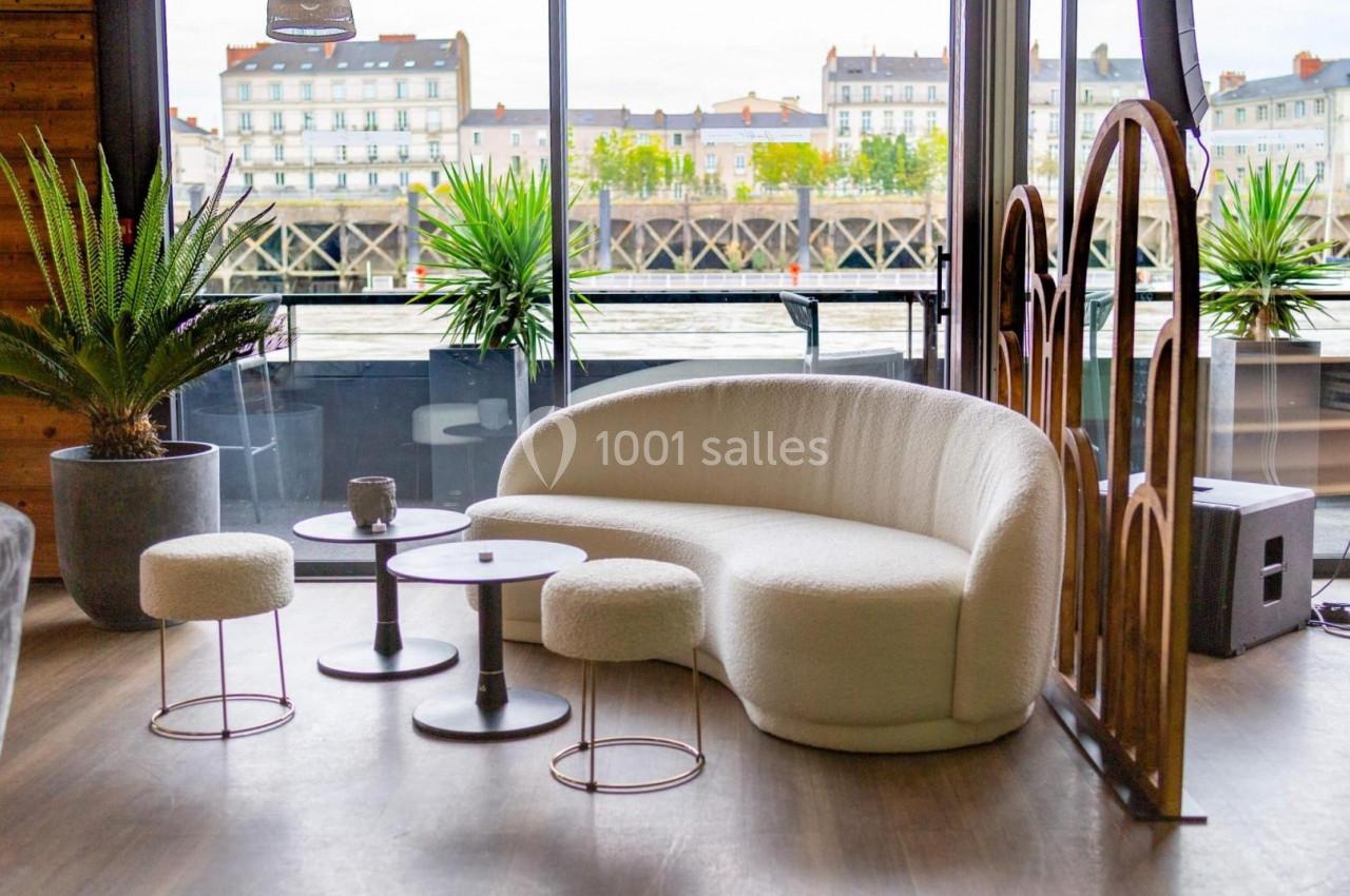 Salon moderne avec canapé blanc incurvé, tables basses rondes, tabourets et vue sur des bâtiments au bord de l'eau.