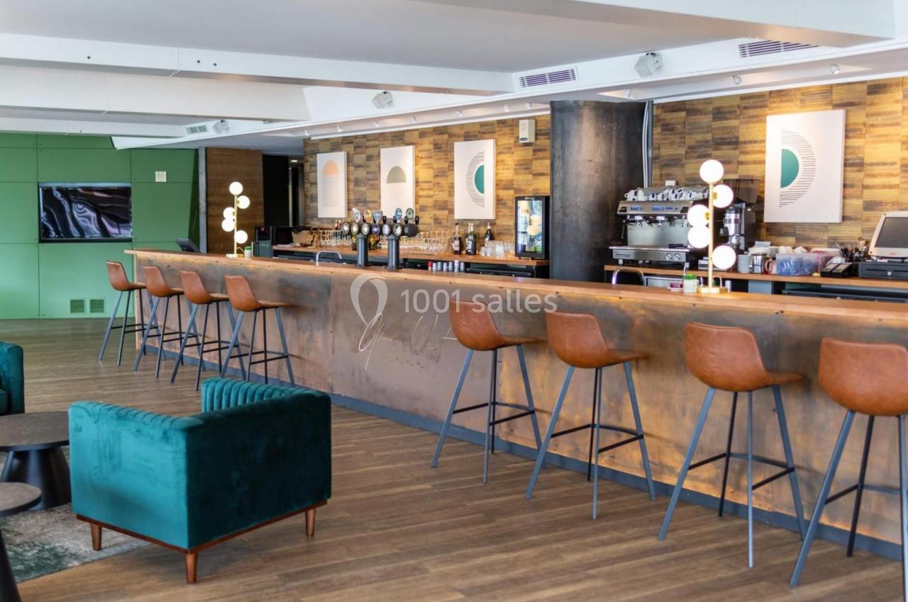 Bar moderne avec comptoir en métal, tabourets en cuir, décor en bois et affiches murales dans un espace lumineux.