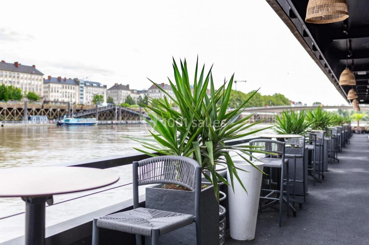 Terrasse avec tables et chaises en bord de rivière, entourée de plantes vertes et vue sur des bâtiments et un pont.