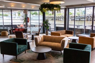 Terrasse avec tables et chaises en bord de rivière, entourée de plantes vertes et vue sur des bâtiments et un pont.