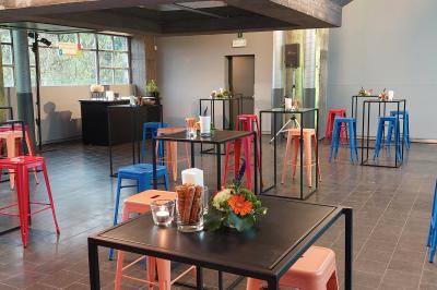 Salle lumineuse avec de grandes baies vitrées, tables dressées avec nappes grises, vaisselle et décoration florale.