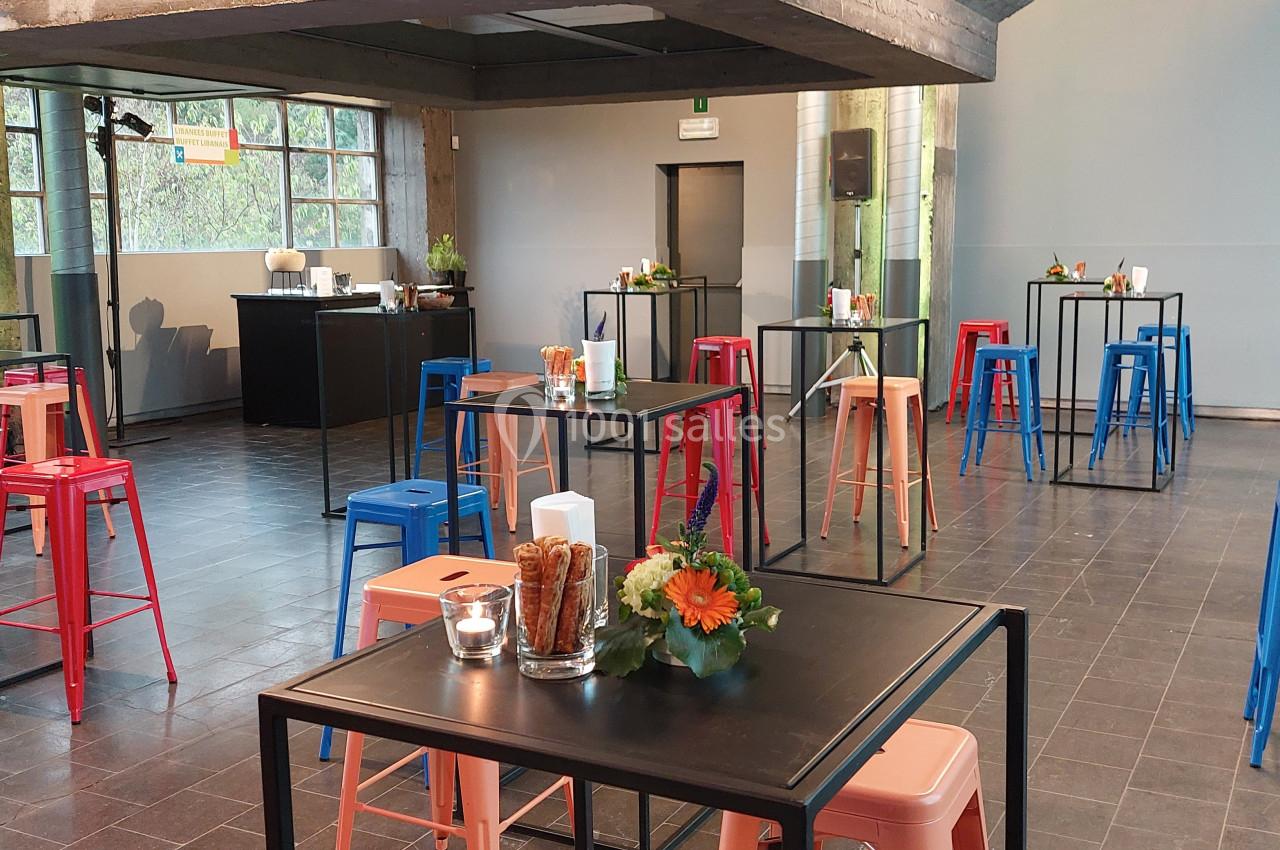 Salle moderne avec tables hautes noires, tabourets colorés et décorations florales, éclairée par des fenêtres.