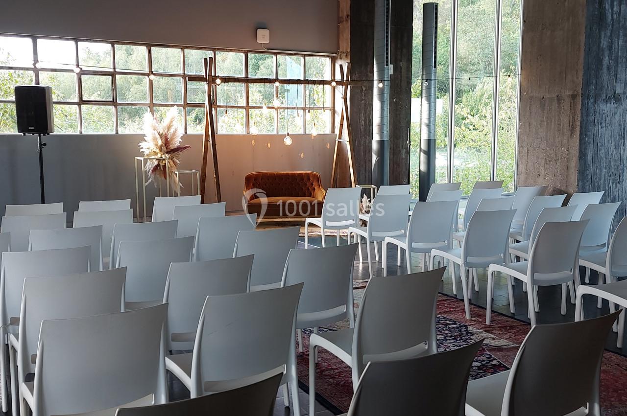 Salle aménagée avec des rangées de chaises blanches, un canapé en fond et de grandes fenêtres laissant entrer la lumière.