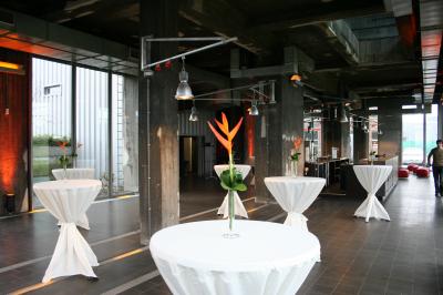 Salle lumineuse avec de grandes baies vitrées, tables dressées avec nappes grises, vaisselle et décoration florale.