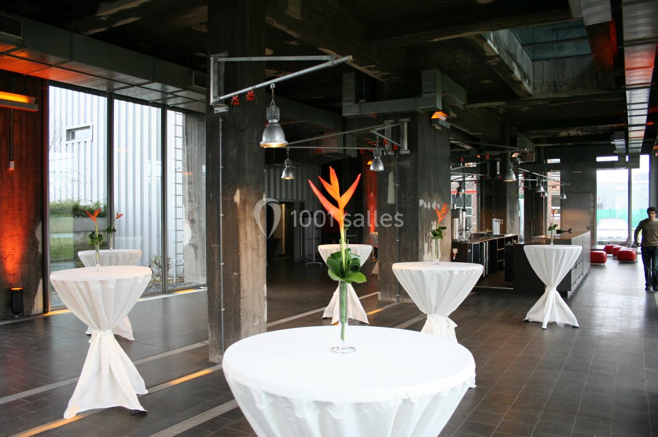 Salle moderne avec tables hautes drapées de blanc, décorées de fleurs tropicales, éclairage industriel.