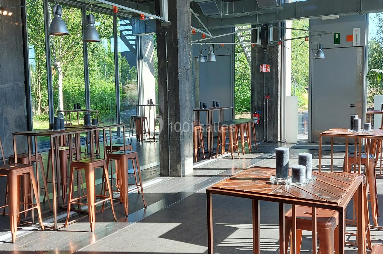 Salle lumineuse avec de grandes baies vitrées, tables hautes en bois et tabourets en métal, vue sur un espace vert.