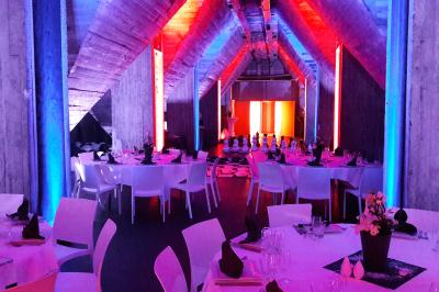 Salle lumineuse avec de grandes baies vitrées, tables dressées avec nappes grises, vaisselle et décoration florale.