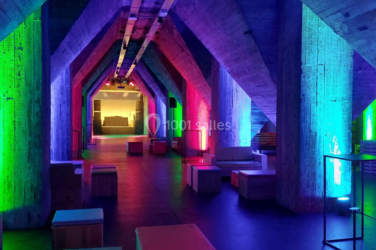 Salle moderne avec piliers en béton illuminés par des lumières colorées, aménagée avec des sièges en bois.