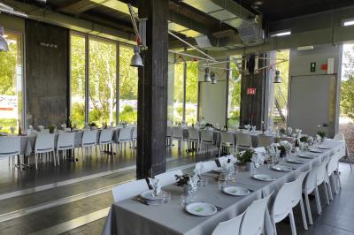 Salle lumineuse avec de grandes baies vitrées, tables dressées avec nappes grises, vaisselle et décoration florale.
