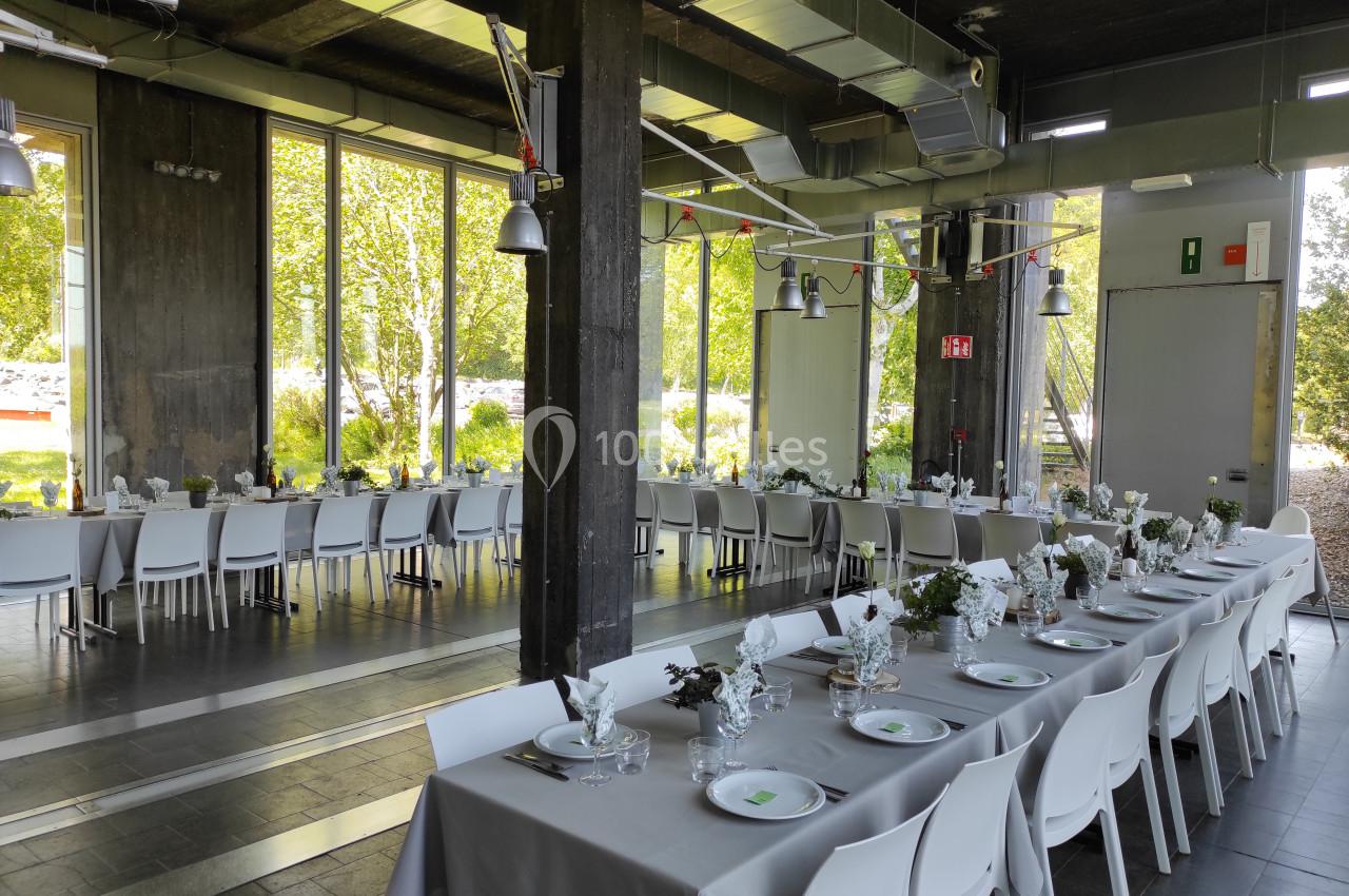 Salle lumineuse avec de grandes baies vitrées, tables dressées avec nappes grises, vaisselle et décoration florale.