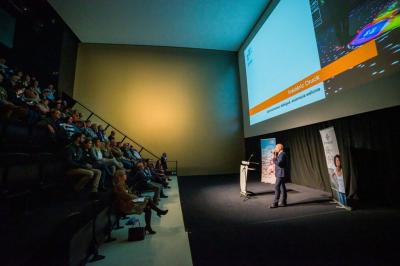 Salle lumineuse avec de grandes baies vitrées, tables dressées avec nappes grises, vaisselle et décoration florale.
