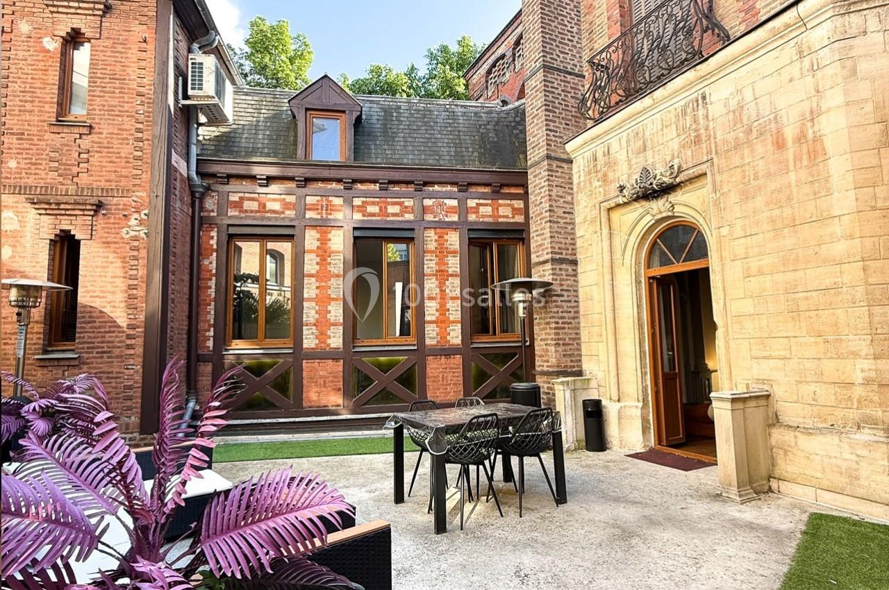 Cour intérieure avec table et chaises, entourée de bâtiments en briques et pierre, et végétation en arrière-plan.